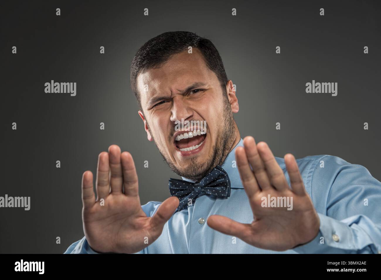 Porträt eines wütenden, verärgerten jungen Mannes in blauem Hemd und Schmetterlingskrawatte mit Händen hoch brüllend isoliert auf grauem Hintergrund. Negative menschliche Emotionen, Hände-Sho Stockfoto