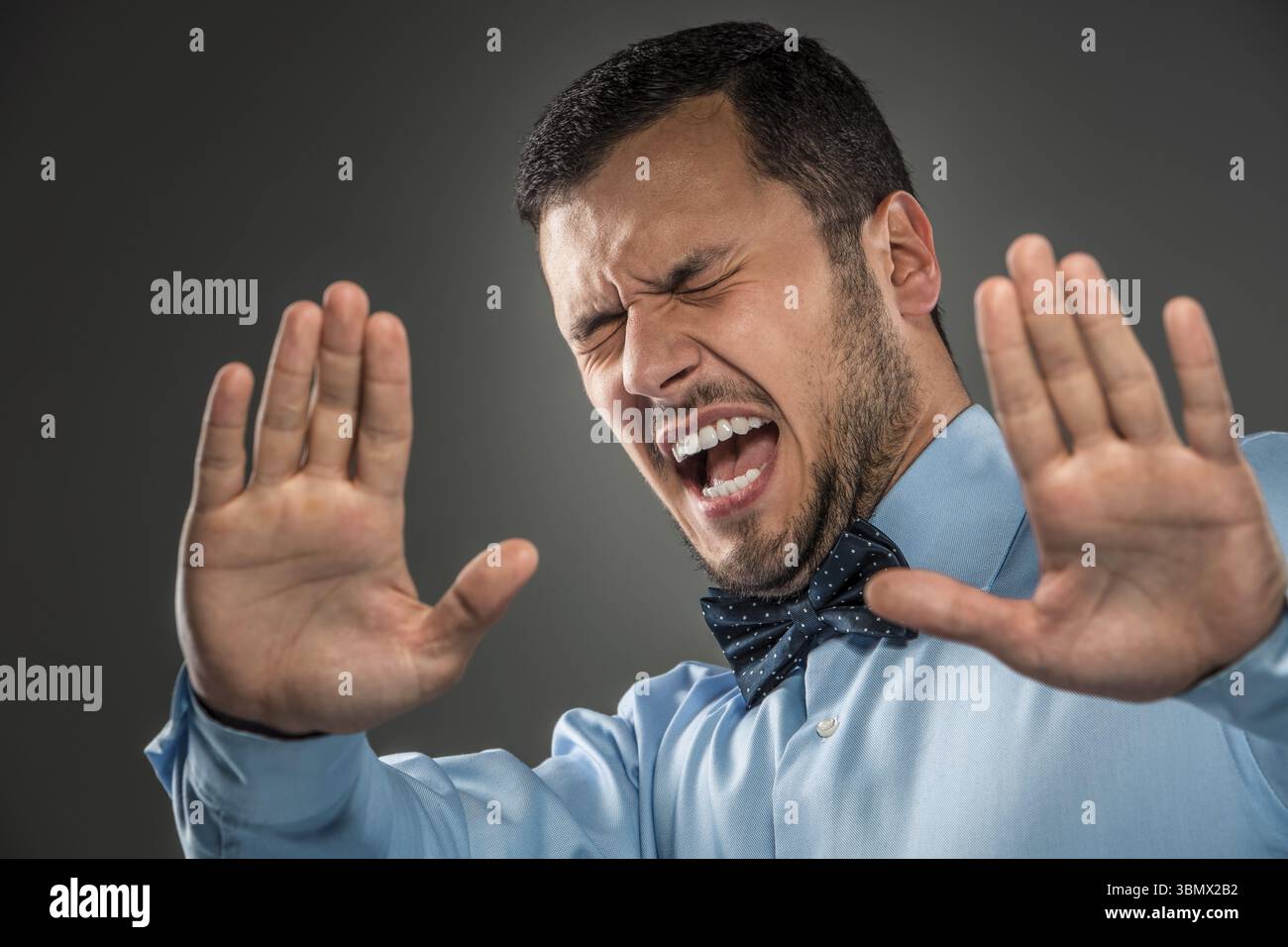 Porträt eines wütenden, verärgerten jungen Mannes in blauem Hemd und Schmetterlingskrawatte mit den Händen hoch schreiend, die Augen geschlossen, isoliert auf grauem Hintergrund. Negatives menschliches Emot Stockfoto