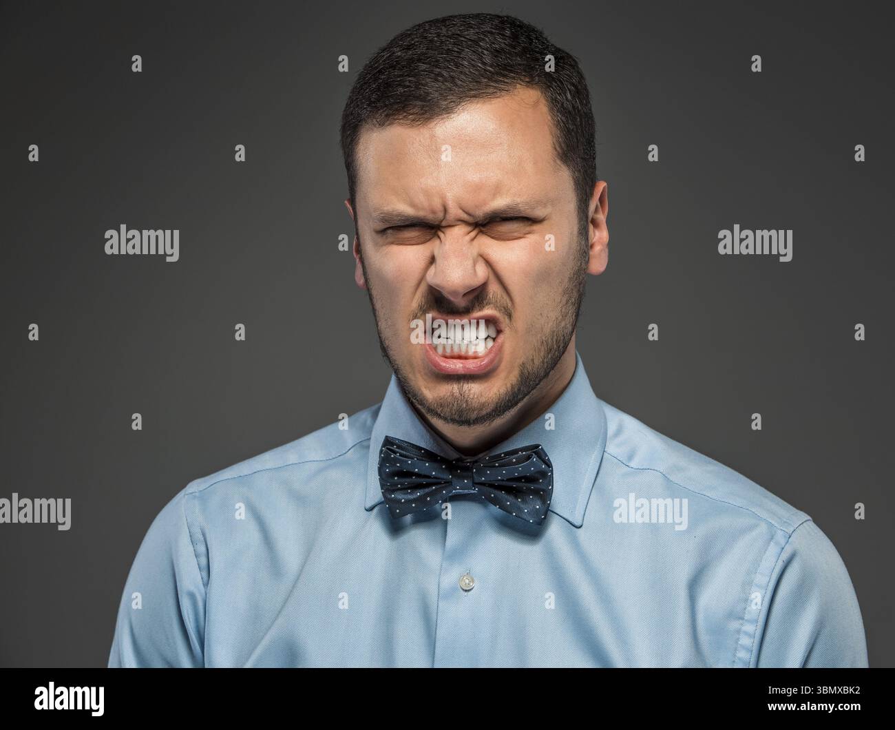 Porträt des wütenden verärgerten jungen Mannes in blauem Hemd und Schmetterlingskrawatte isoliert auf grauem Studiohintergrund. Negative menschliche Emotion, Gesichtsausdruck. Schließen Stockfoto