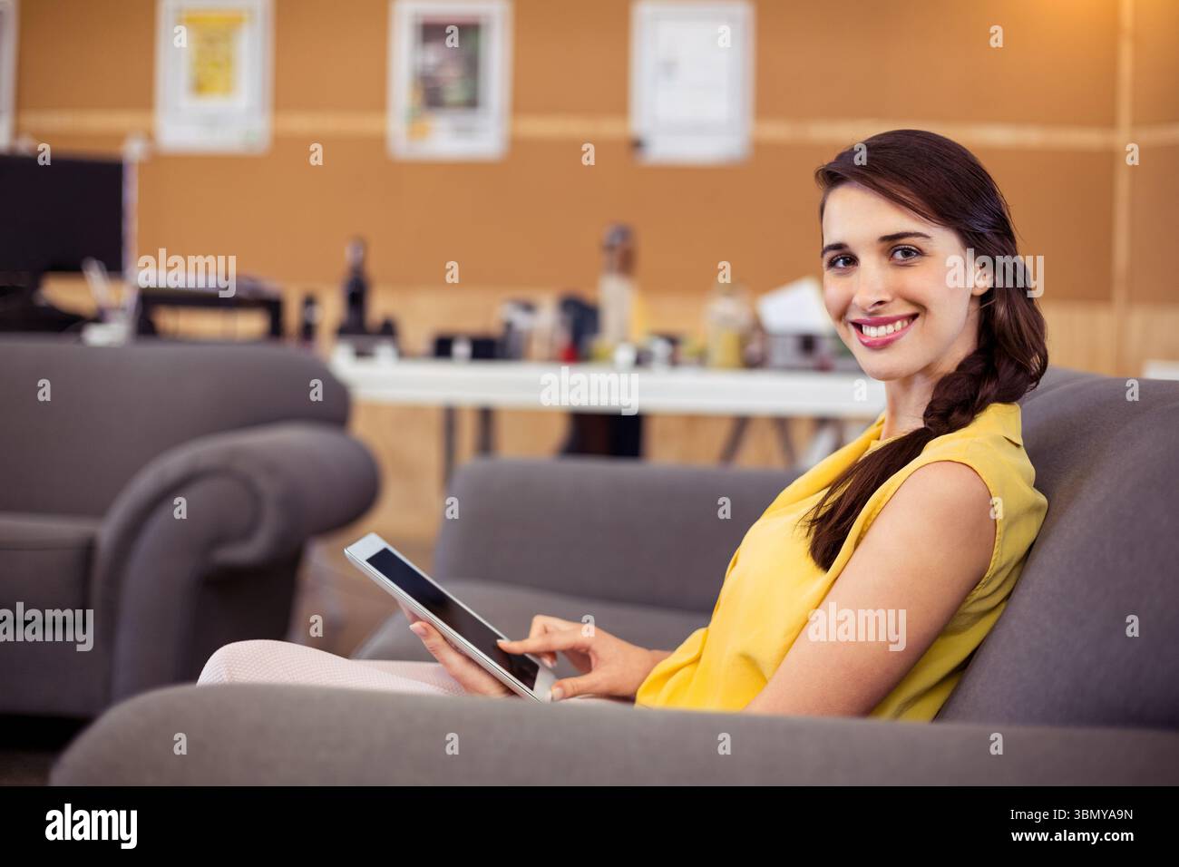 Erwachsene Frau, die auf grauem Sofa in der Bürolounge sitzt, Senfbluse mit Tablet und Kopierraum trägt Stockfoto