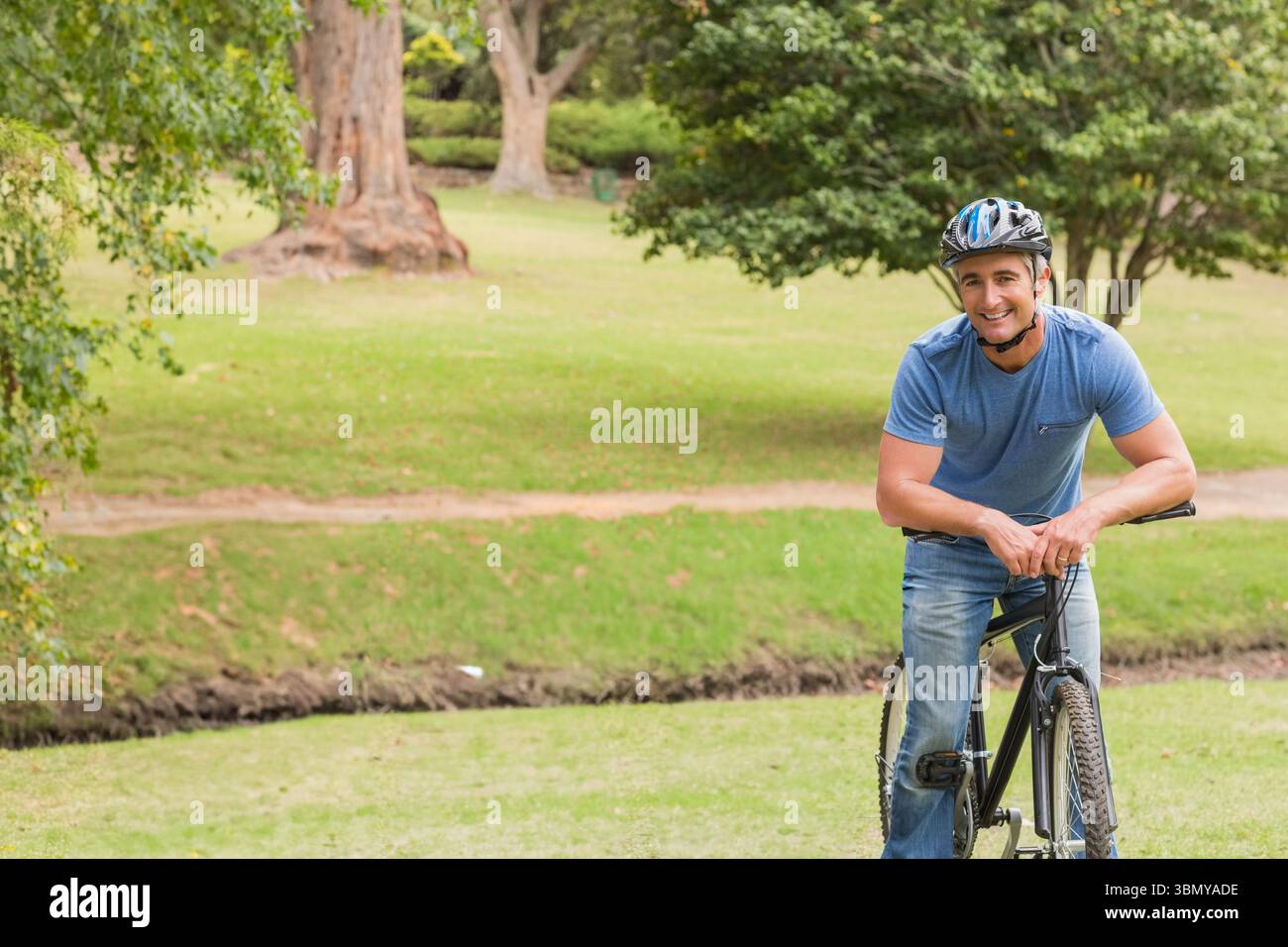 Mann, der sich auf ein schwarzes Fahrrad stützt, trägt einen mehrfarbigen Helm in der Nähe des Wanderweges im Park, Kopierraum Stockfoto