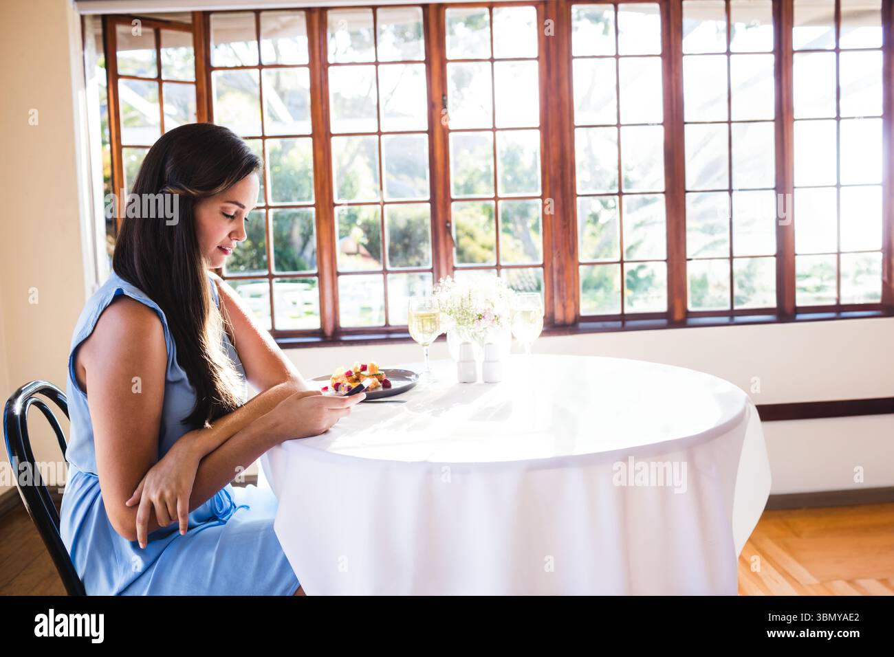 Frau in ärmellosem blauem Kleid, die ihr Smartphone am Esstisch zu Hause mit geschnittenem Obst überprüft Stockfoto