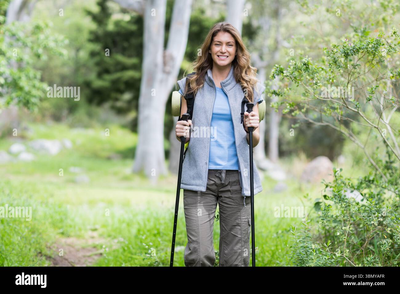 Frau, die durch grasbewachsene Waldlichtung wandert, trägt eine Weste, packt Wanderstöcke und trägt einen Rucksack Stockfoto