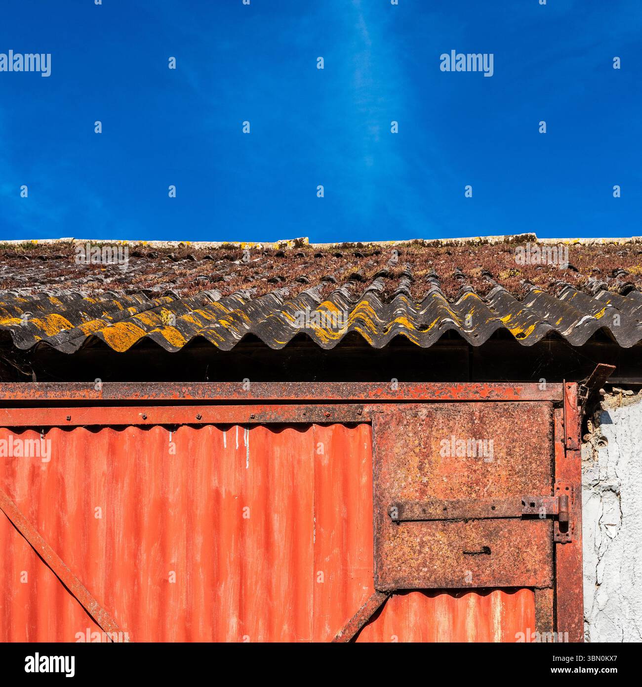 Alte, rostrote Metalltür und verwittertes Fabrikdach vor blauem Himmel. Abstrakt. Texturen. Kontraste. Stockfoto