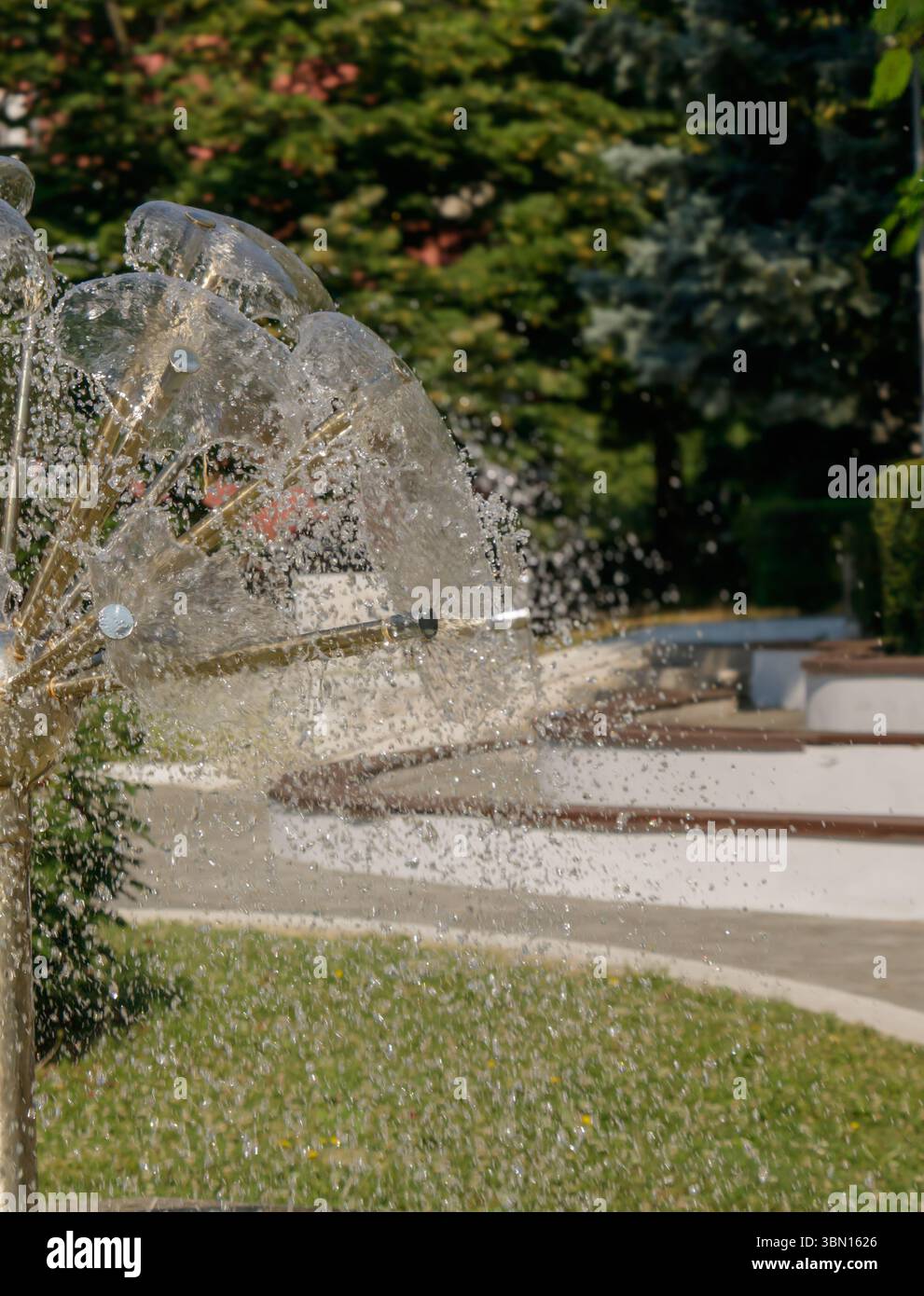 Helles Sonnenlicht beleuchtet einen einzigartigen Wasserbrunnen in einem öffentlichen Park. Der Springbrunnen sprüht Wasser in einem künstlerischen Muster, während üppiges Grün umgeben ist Stockfoto