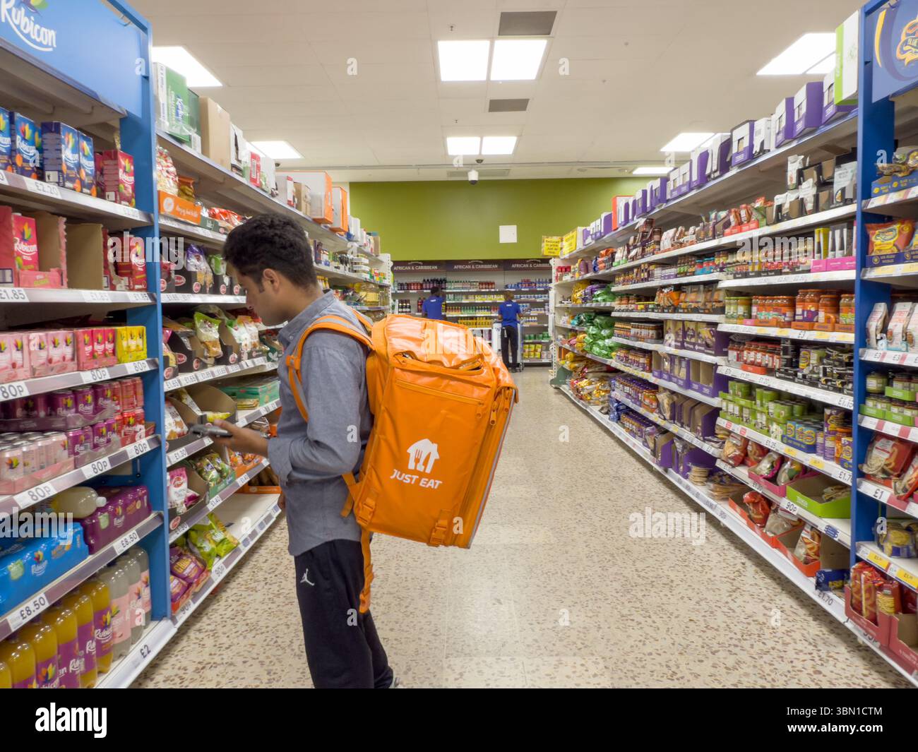 In einem Supermarktladen laden laden ein Just-Eat-Shopper und ein Lieferfahrer mit orangefarbenem Rucksack im Londoner Supermarkt zur Kundenbestellung ein Stockfoto
