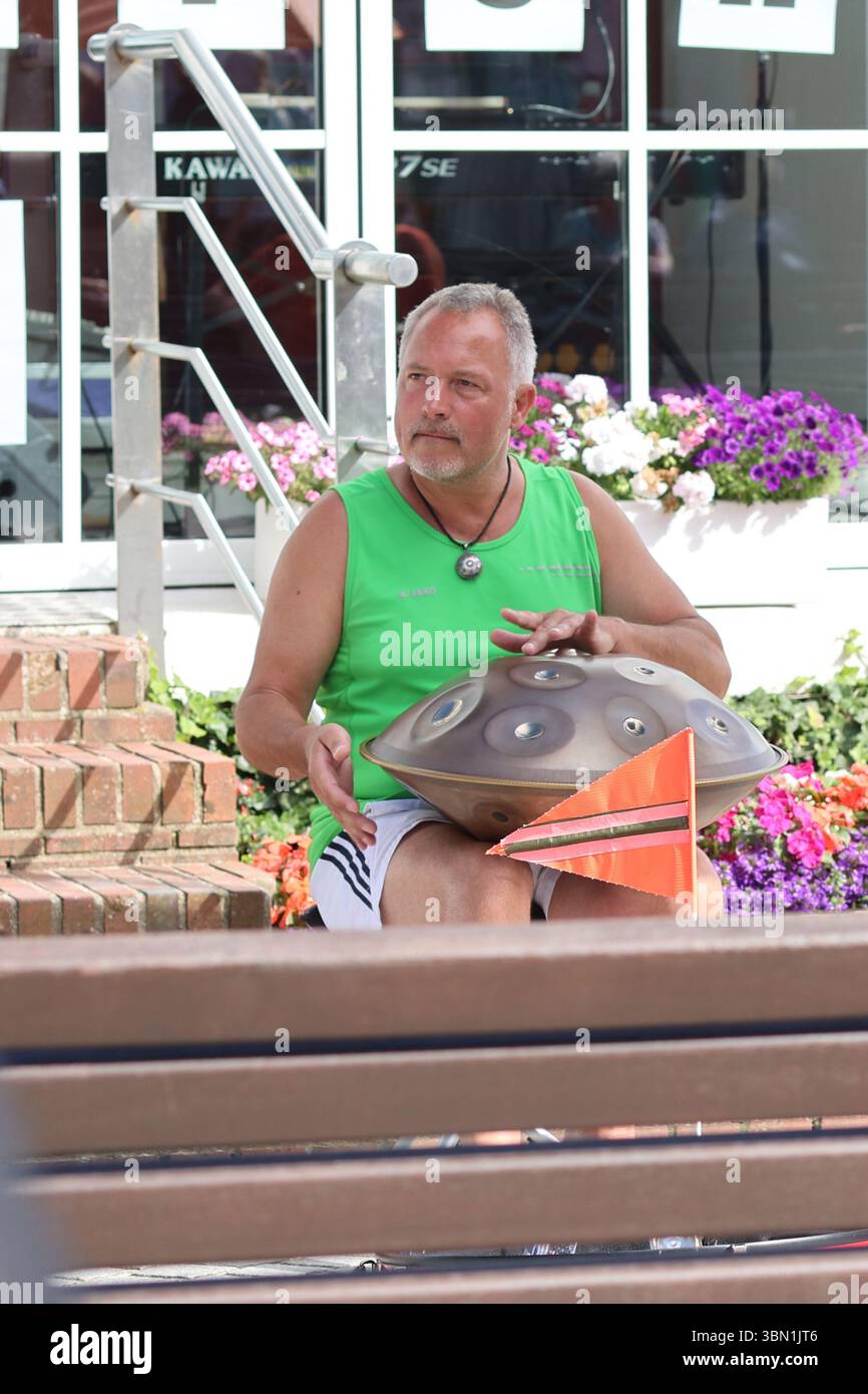 Amrum, Deutschland - 5. August 2024: Ein Mann in einem grünen Tanktop spielt auf einer Handpfanne und fesselt Passanten mit melodischen Klängen, während er darauf sitzt Stockfoto