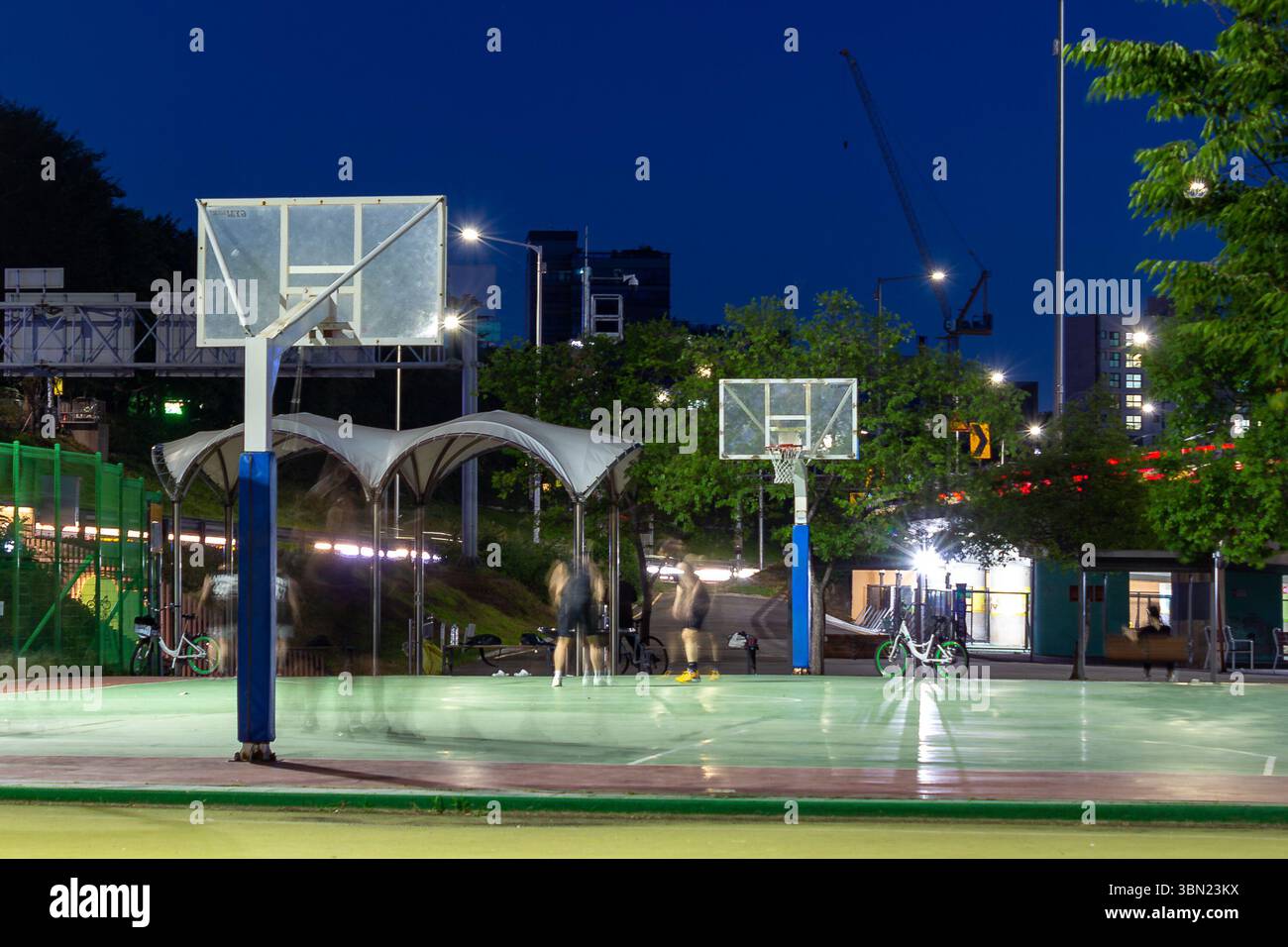 Nächtlicher Blick auf einen öffentlichen Basketballplatz mit Spielern in Bewegung, beleuchtet von Straßenlaternen, vor dem Hintergrund von städtischen Gebäuden und einem Dee Stockfoto