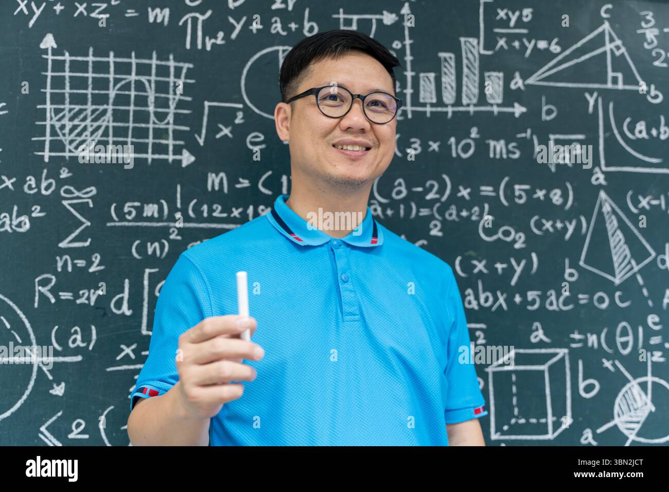 Porträt eines lächelnden asiatischen Lehrers in blauem Hemd, der Kreide vor einer Tafel mit mathematischen Formeln hält. Perfekt für den Bildungsbereich Stockfoto