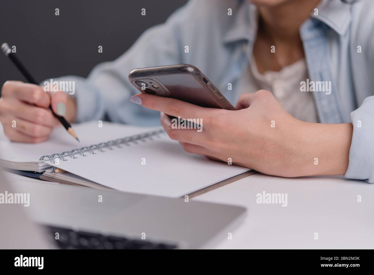 Junge Frau, die ein Smartphone benutzt, während sie Notizen macht und dabei die Balance zwischen digitaler Kommunikation und traditioneller Notiz in einem modernen Worksp zeigt Stockfoto