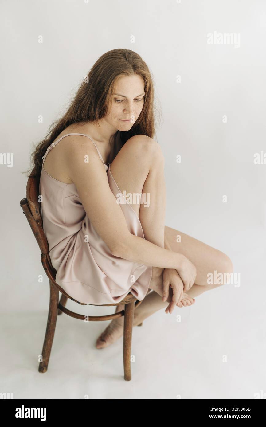 Rote, langhaarige, Traurige Frau sitzt auf einem Stuhl vor einem weissen Hintergrund Stockfoto