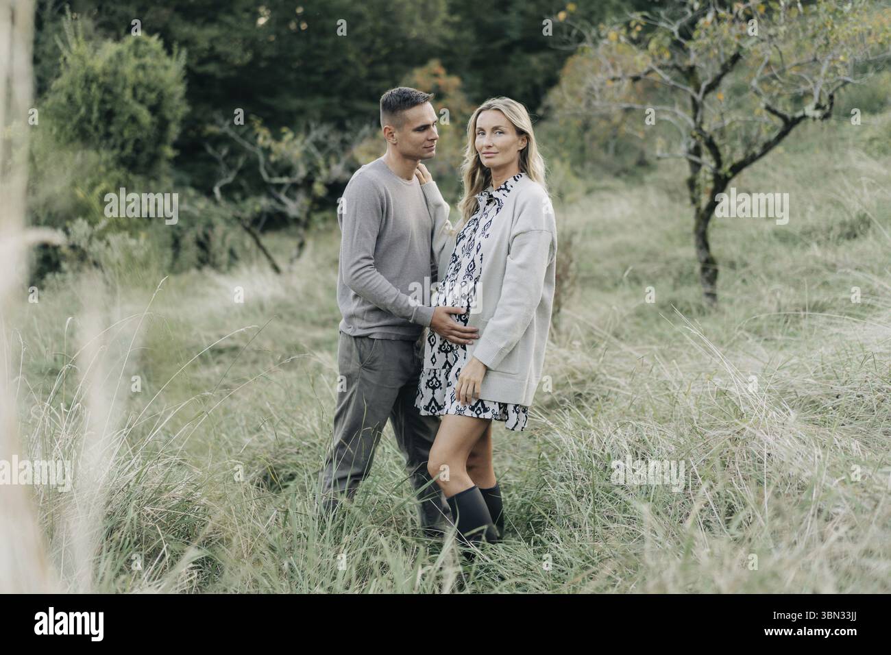Glueckliches schwangeres Paar laechelnd und umarmend bei Sonnenuntergang zusammen im Garten Stockfoto