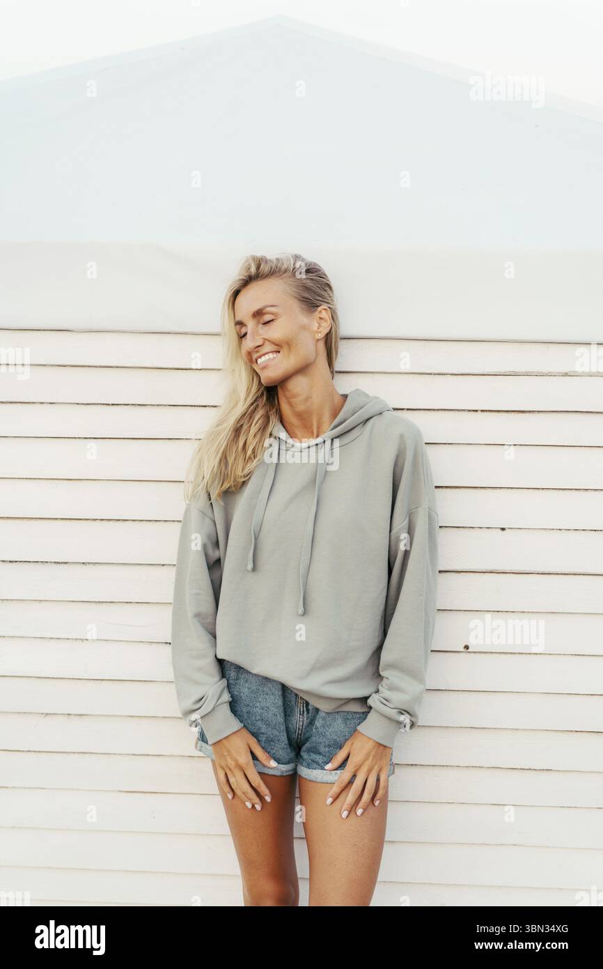 Niedliche schöne gebräunte blonde Frau in Jeansshorts und einem stylischen Hoodie, die Spaß hat und aufrichtig lacht Stockfoto