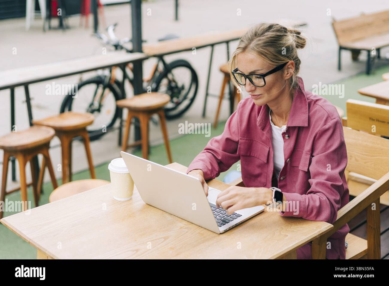 Junge blonde Geschaeftsfrau sitzt auf der Terrasse eines Cafes und arbeitet an einem Lapto Stockfoto