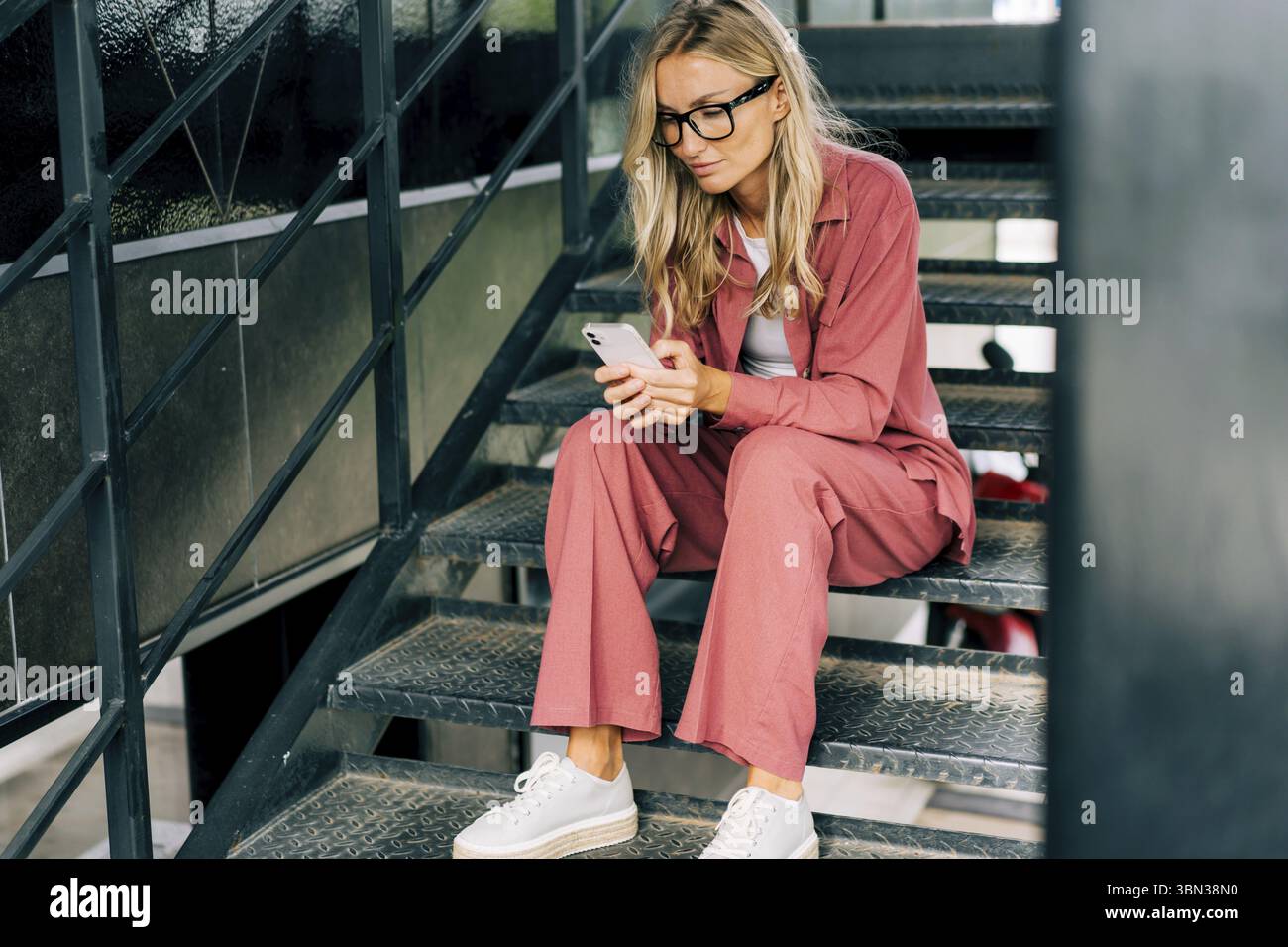 Junge laessige Frau, die ihr Smartphone benutzt, waehrend sie auf den Eisenstufen eines modernen Buerogebaeudes sitzt Stockfoto