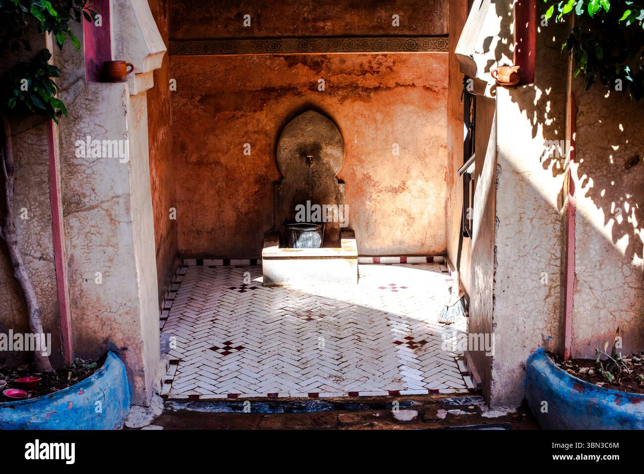 Nische mit Brunnen und Kupferbecken mit alten orangen Wänden und weißem geometrischem Backsteinboden zwischen Bäumen in den Straßen von Marrakesch Marokko Stockfoto