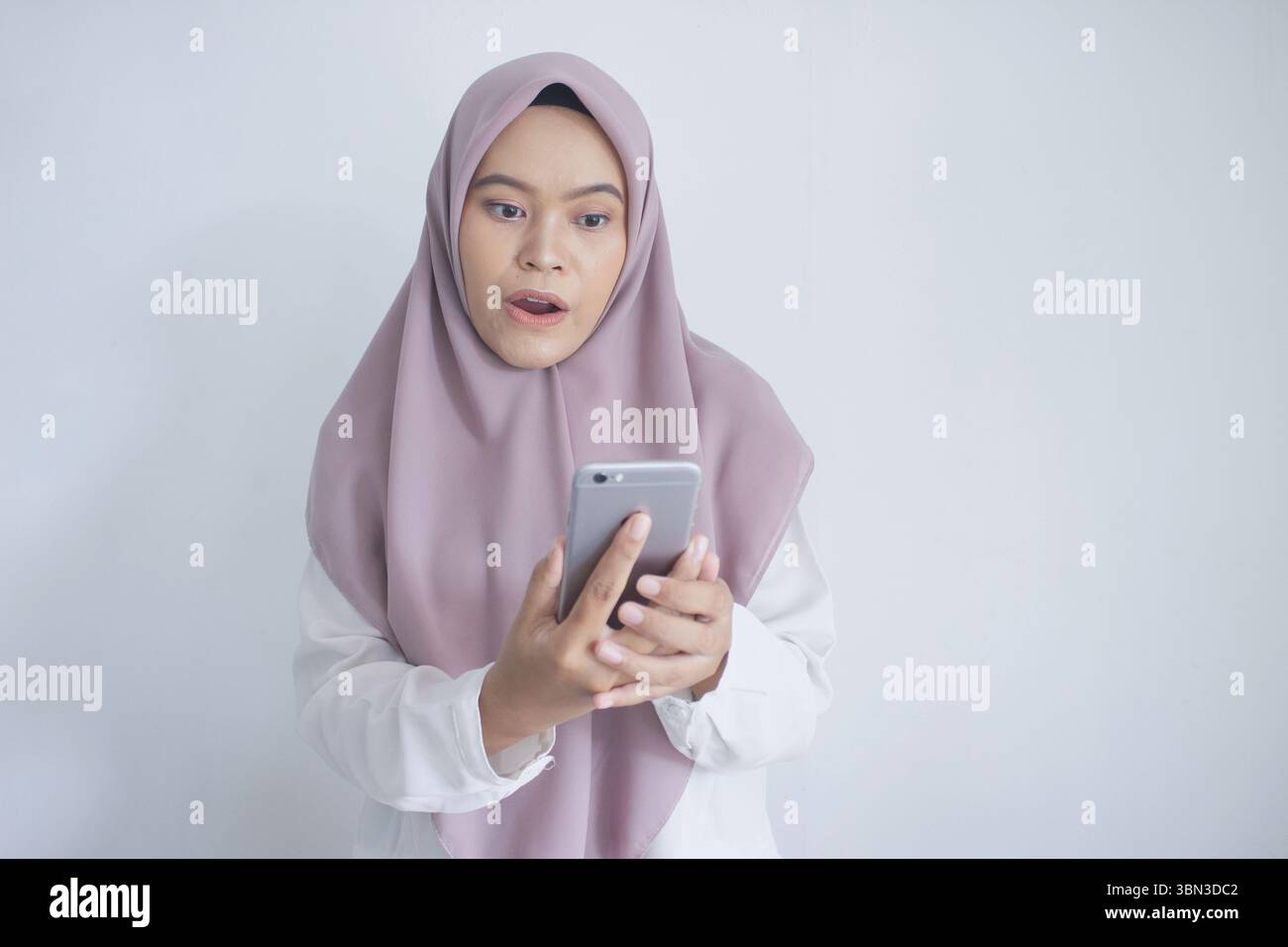 Die junge muslimische asiatin trägt einen violetten Hijab und hält ein Handy. Sie ist von etwas am Telefon überrascht oder schockiert Stockfoto