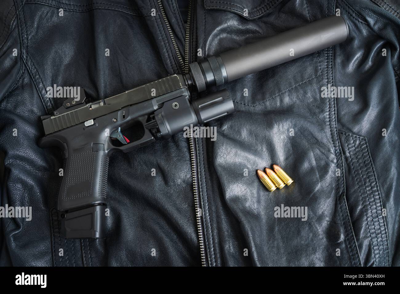 Eine taktische Pistole mit Schalldämpfer, rotem Punkt und Taschenlampe liegt auf einer schwarzen Lederjacke neben Munition. Stockfoto