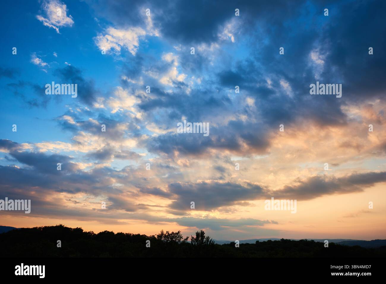 Atemberaubender Himmel voller dramatischer Wolken übergeht bei Sonnenuntergang von tiefem Blau zu sanften Pastelltönen. Horizont gesäumt von silbrigen Hügeln und Wäldern, die der ruhigen und weitläufigen Landschaft Tiefe verleihen. Stockfoto