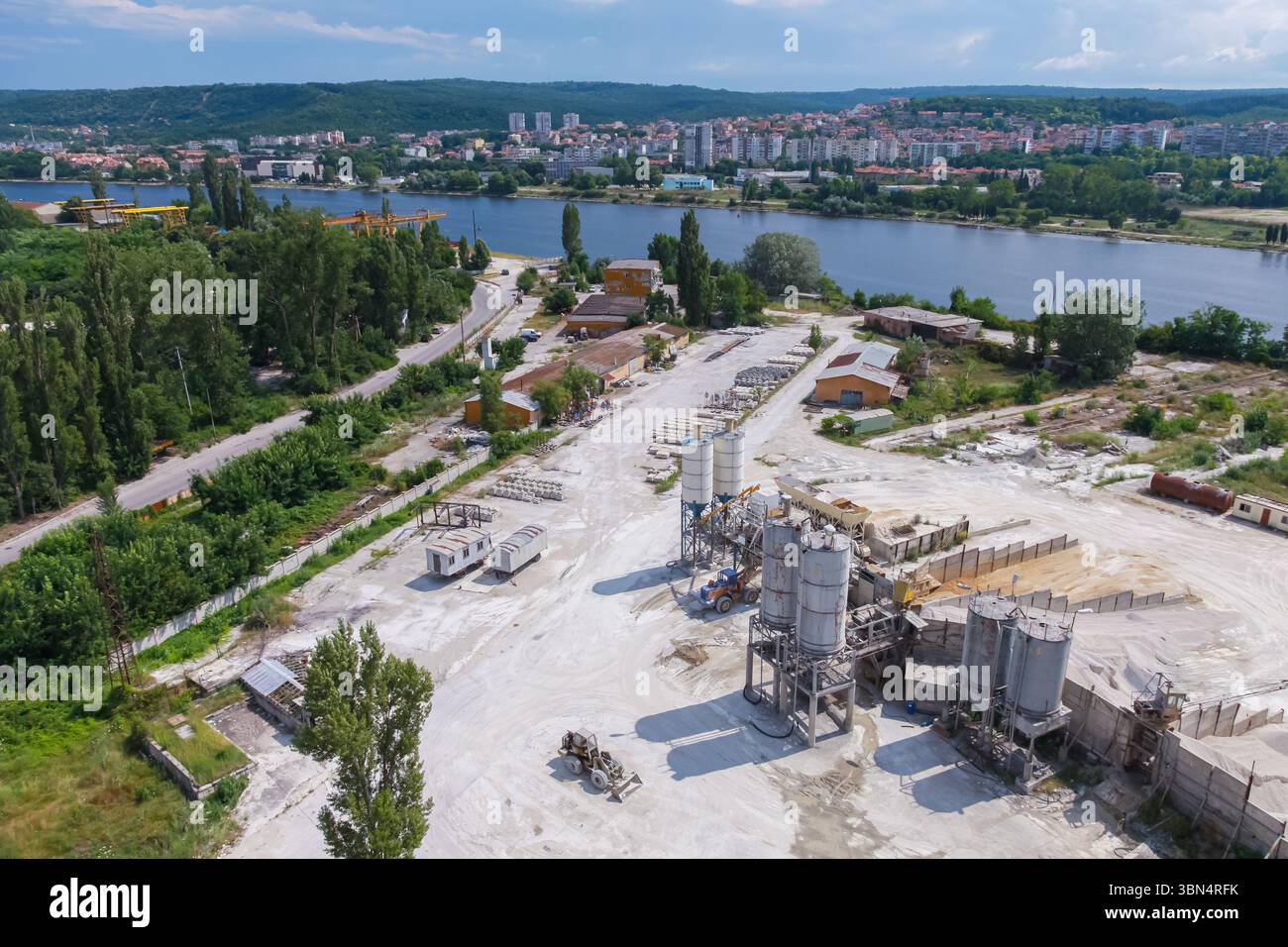 Aus der Vogelperspektive ist eine weitläufige Industrieanlage, umgeben von grünen Bäumen, neben einem ruhigen Fluss in der Nähe von Wohngebieten zu sehen. Varna, Bulgari Stockfoto