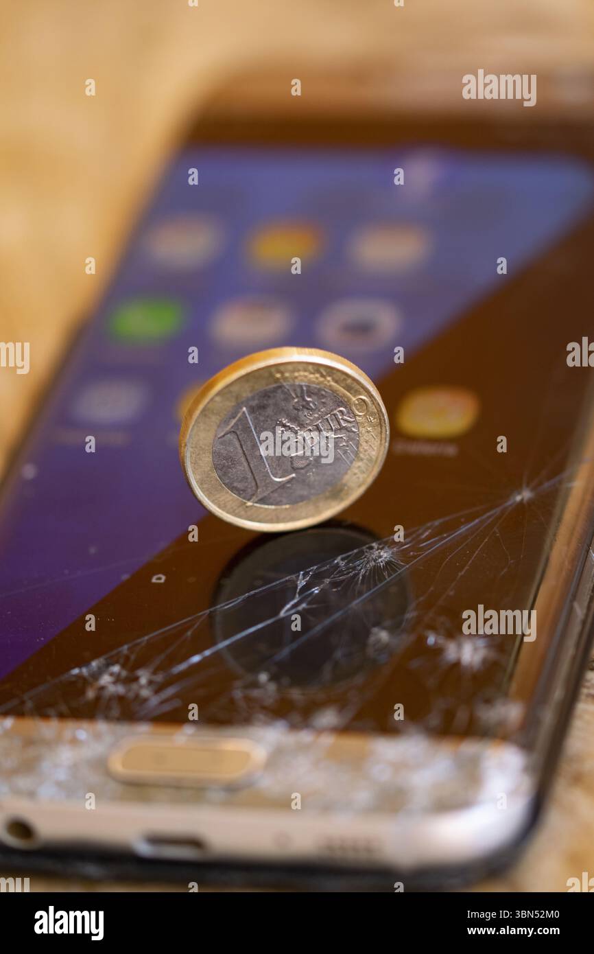 Euro-Münze auf einem zerrissenen Smartphone-Bildschirm, die wirtschaftlichen Wert im Vergleich zu zerbrechlicher oder veralteter Technologie symbolisiert. Digital Decay Conep Stockfoto