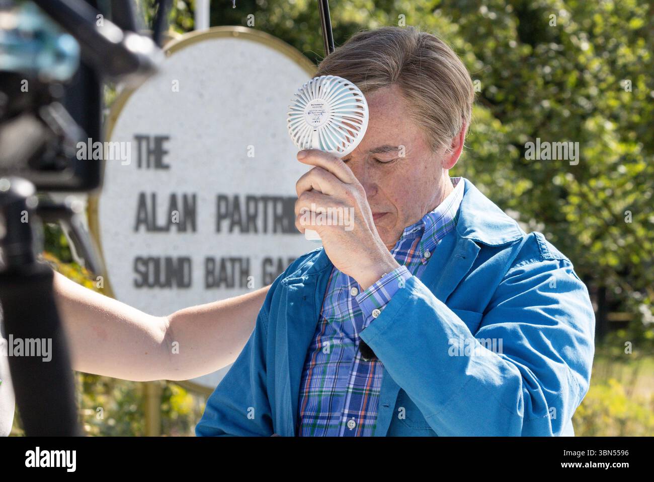 Hampton Court Palace, Surrey, Großbritannien. 30. Juni 2025. Steve Coogan, gekleidet als sein Charakter Alan Partridge, filmt im Alan Partridge Sound Bath Garden ein Stück zu Kameras, wobei er einen Ventilator und einen Regenschirm verwendet, um zwischendurch die heißen Temperaturen abzukühlen. Prominente und Pressebesucher genießen das Blumenfest. Wunderschöne Gartendesigns, Produkte und Blumendekorationen werden vom 1. Bis 6. Juli auf dem HAMPTON Court Palace Garden Festival gezeigt. Quelle: Imageplotter/Alamy Live News Stockfoto