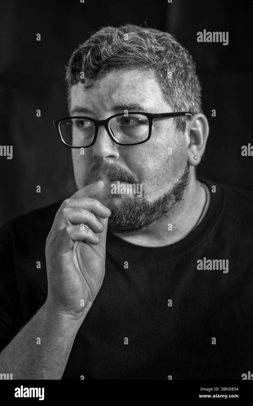 Studio-Porträt eines jungen kaukasischen Mannes, der eine elektronische Zigarette raucht und Brille und Ohrring trägt, mit Bart und Schnurrbart auf schwarzem Hintergrund Stockfoto