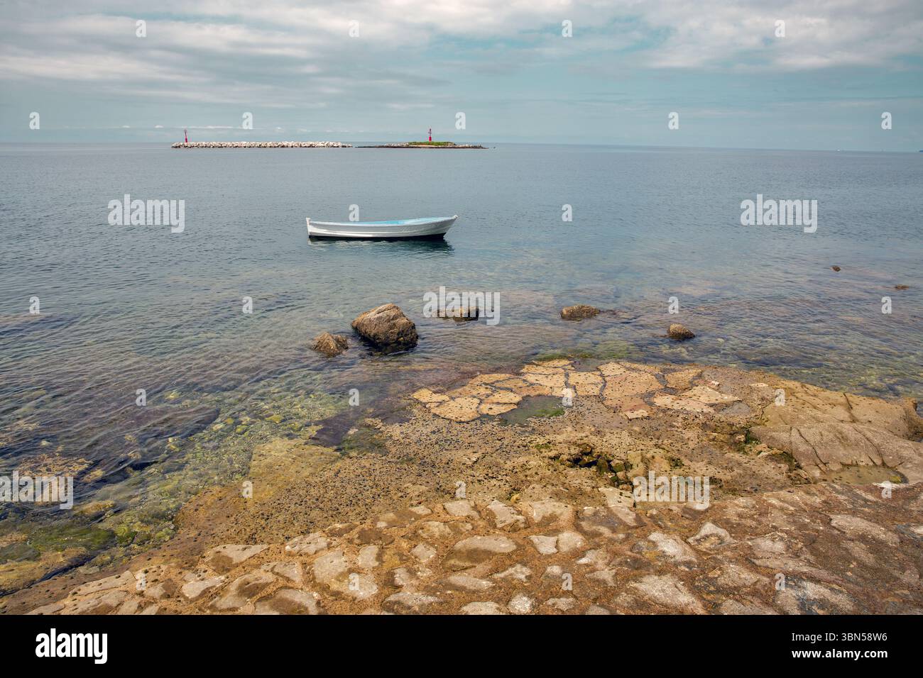 Kleines weißes Boot, das auf ruhigem Meer in der Nähe der felsigen Küste schwimmt. Leuchtturm auf Barbaran Island im Hintergrund. Friedliche Küstenlandschaft, klares Wasser, Sommer t Stockfoto