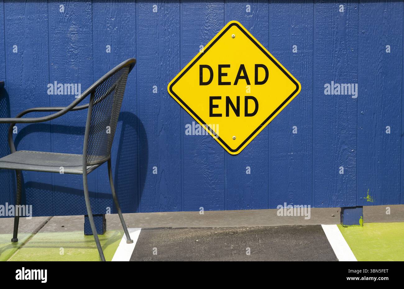 Ein dunkler Metallstuhl sitzt leise neben einem auffälligen gelben „Sackgasse“-Schild vor einer leuchtend blauen Wand – eine unerwartete, poetische urbane Szene Stockfoto