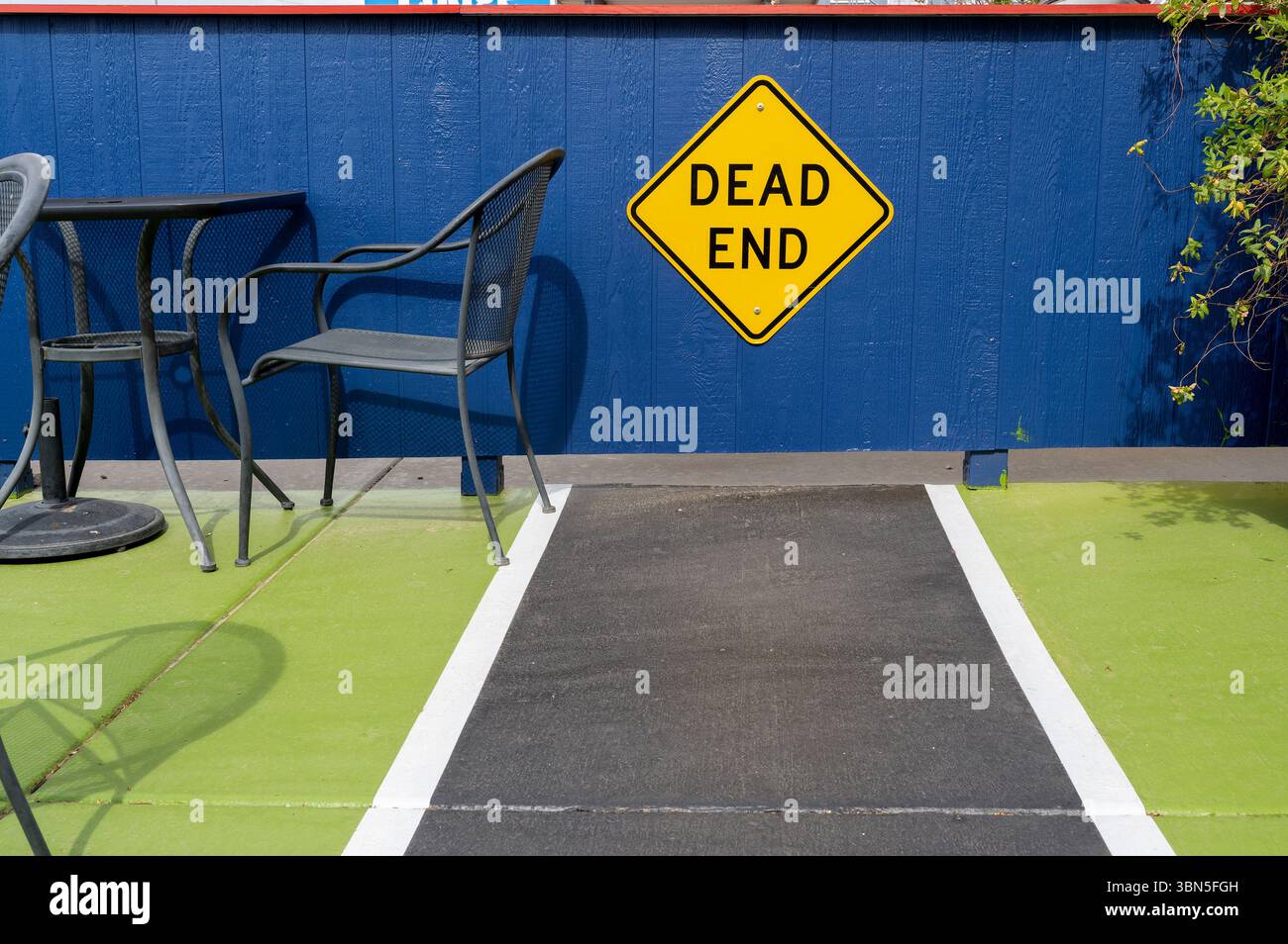 Ein dunkler Metallstuhl sitzt leise neben einem auffälligen gelben „Sackgasse“-Schild vor einer leuchtend blauen Wand – eine unerwartete, poetische urbane Szene Stockfoto