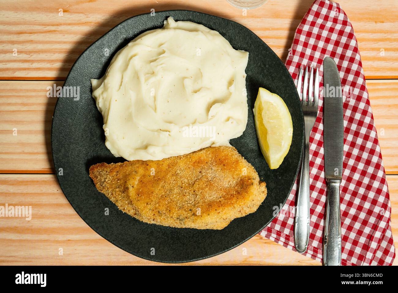 Oben ein paniertes Fischfilet serviert mit Kartoffelpüree und einer Zitronenscheibe auf einem schwarzen Teller auf einem Holztisch Stockfoto