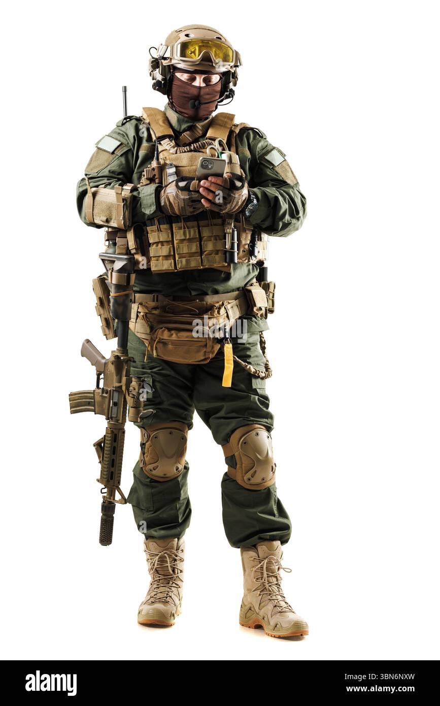 Ein Soldat in voller taktischer Ausrüstung und Helm benutzt ein mobiles Gerät, während er mit einem Gewehr bewaffnet ist. Zeigt moderne militärische Ausrüstung, Einsatzbereitschaft und Te Stockfoto