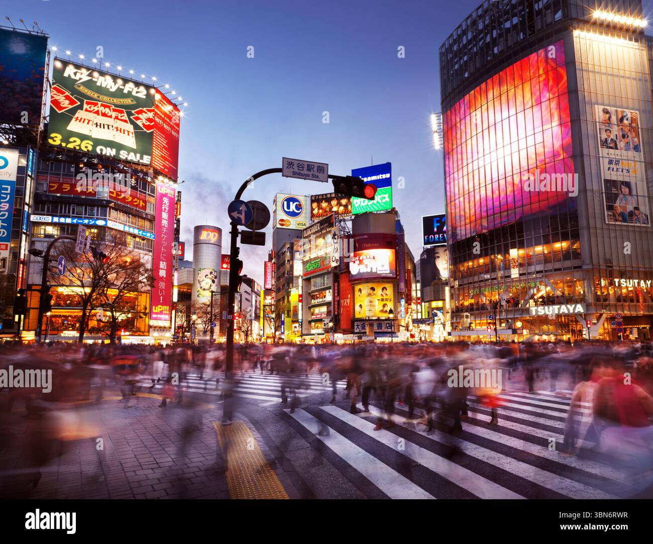 Menschenmenge überquert die Meiji dori Straße am Bahnhof Shibuya, beleuchtet mit bunten Schildern während Sonnenuntergang. Shibuya, Tokio, Japan. Künstlerische Bewegungsunschärfe Stockfoto
