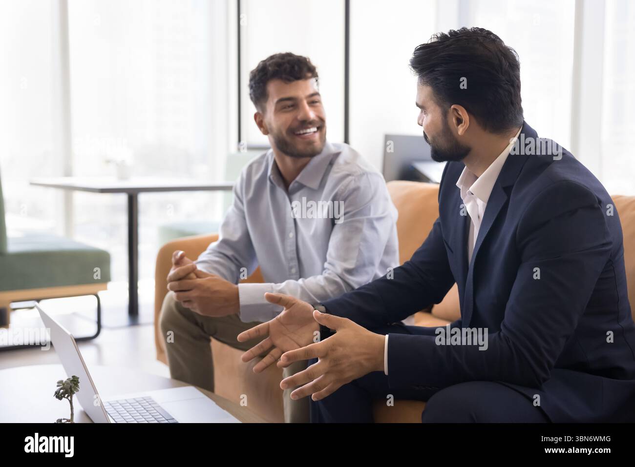 Ein lächelnder junger Geschäftsmann spricht mit einem männlichen Kollegen, der auf der Couch sitzt Stockfoto
