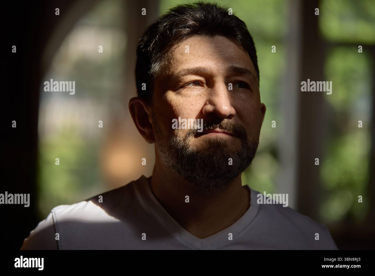 Emotionale Tiefe. Besinnlicher Mann, der in sanftem Fensterlicht steht, mit nachdenklichem Ausdruck Stockfoto