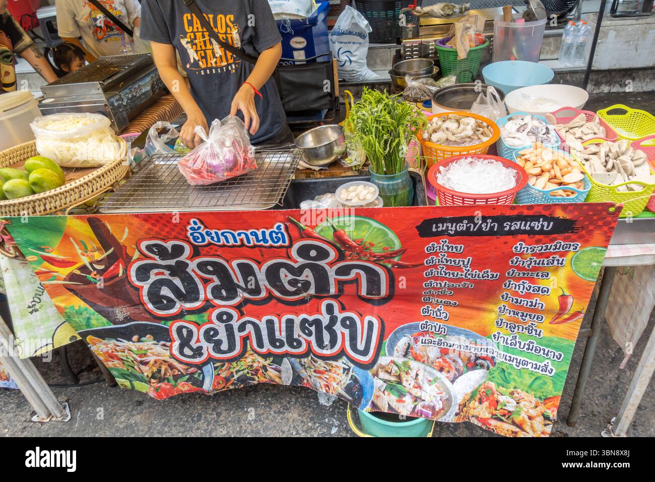 Ein Lebensmittelverkäufer steht hinter einem Verkaufsstand, der „Som Tum & Yum Zaap“ (Papaya-Salat und scharfer Salat) wirbt. Pratunam Markt, Ratchathewi, Bangkok Stockfoto