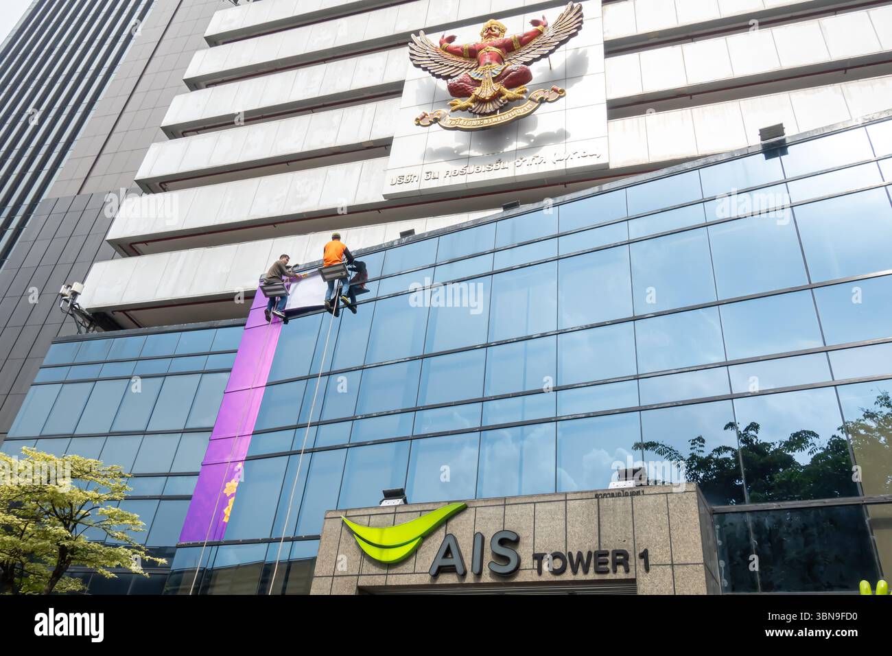 Arbeiter reinigen die Fenster eines Gebäudes, das mit einem großen Adleremblem geschmückt ist. Garuda, Emblem von Thailand, AIS Tower 1 (5G), Bangkok, Thailand Stockfoto