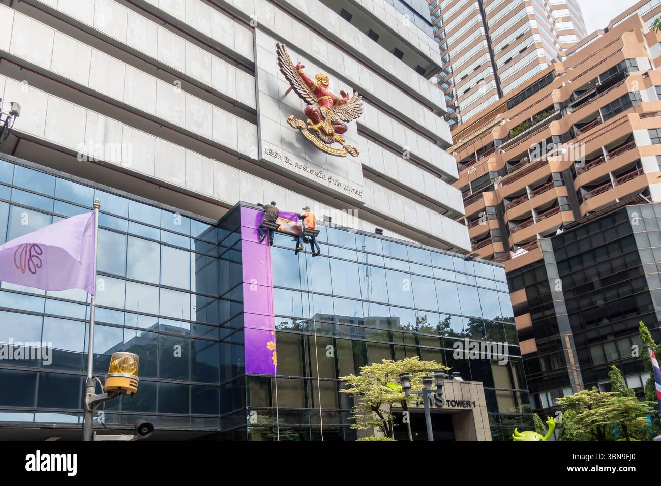 Arbeiter reinigen die Fenster eines Gebäudes, das mit einem großen Adleremblem geschmückt ist. Garuda, Emblem von Thailand, AIS Tower 1 (5G), Bangkok, Thailand Stockfoto
