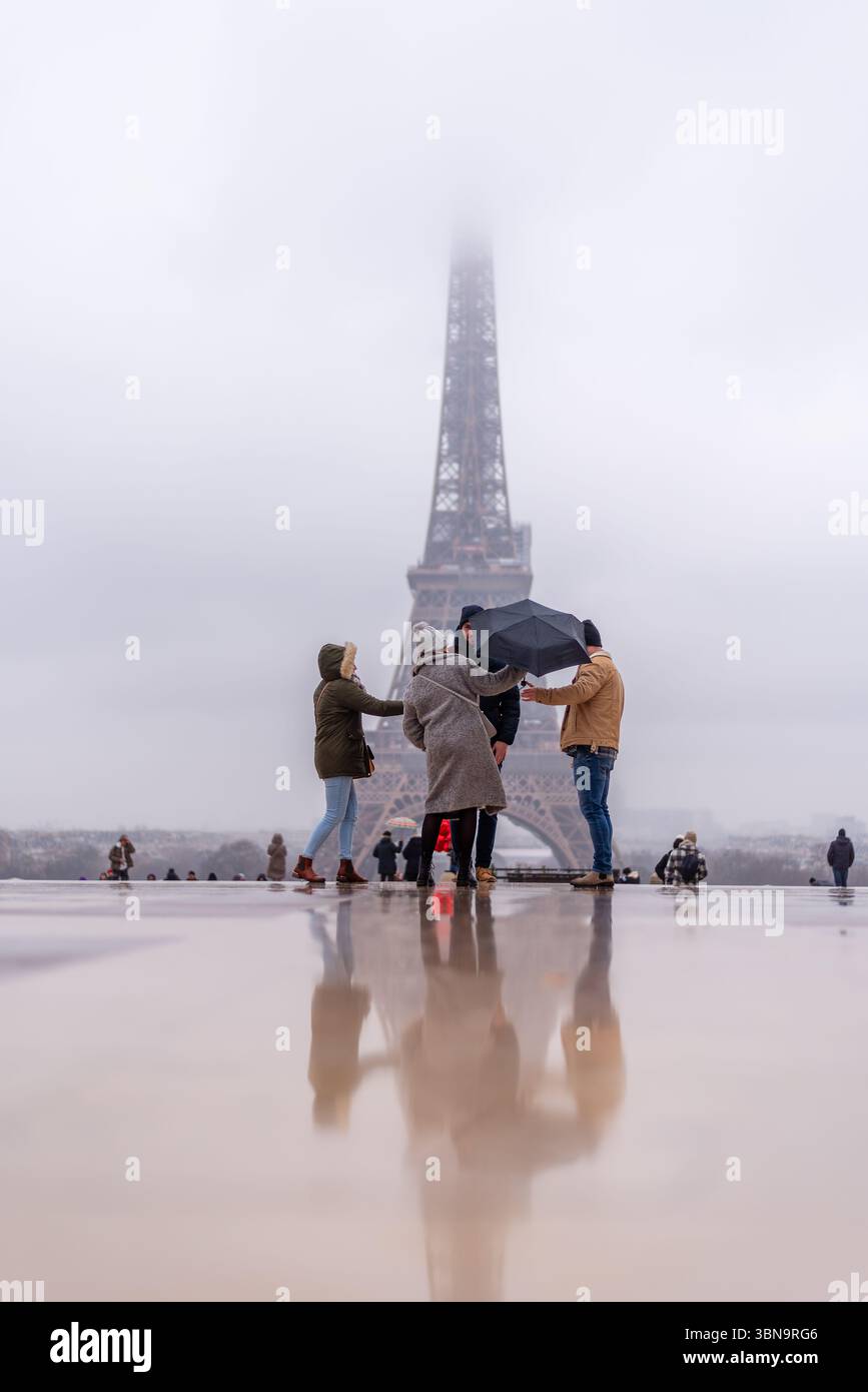 Touristen mit einem Sonnenschirm an einem sehr regnerischen und bewölkten Tag vor dem Eiffelturm, der teilweise von den niedrigen hängenden Wolken verborgen ist. Moody Wetter Stockfoto
