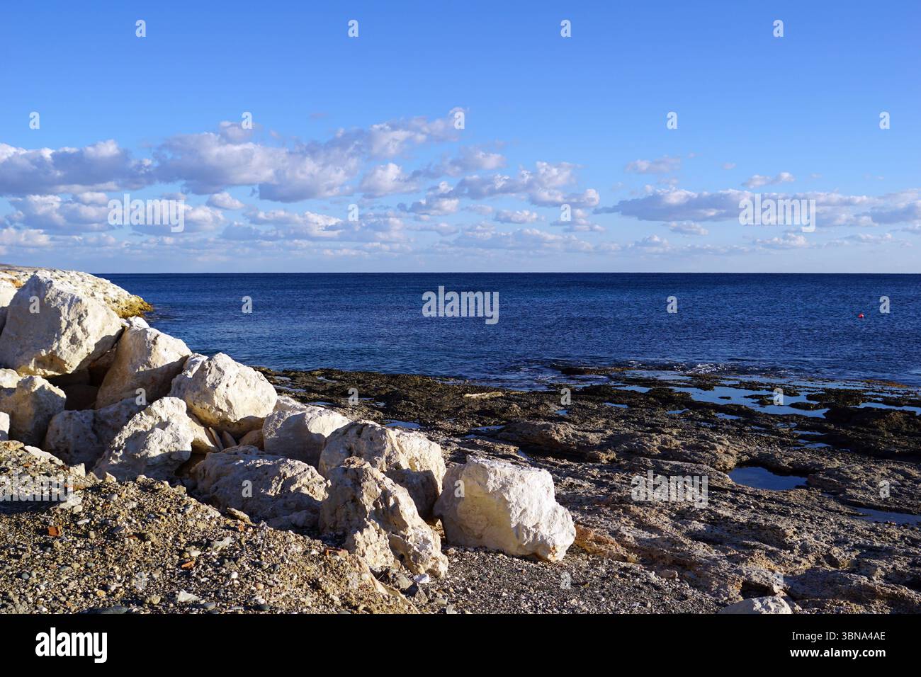 Eine felsige Küste mit einem klaren blauen Himmel und verstreuten weißen Wolken. Das felsige Ufer besteht hauptsächlich aus weißen und hellgrauen Felsen, wobei einige dunklere Felsen durchsetzt sind. Ein kleines, dunkles Objekt, möglicherweise eine Boje, ist im tiefblauen Ozean sichtbar. Der Ozean erstreckt sich bis zum Horizont und trifft den Himmel am fernen Punkt. Die Perspektive des Bildes ist vom Ufer aus und blickt auf die Weite des Ozeans. Ein Künstlerauge und fantasievolle Bildunterschrift. Die Sehenswürdigkeiten Zyperns Stockfoto