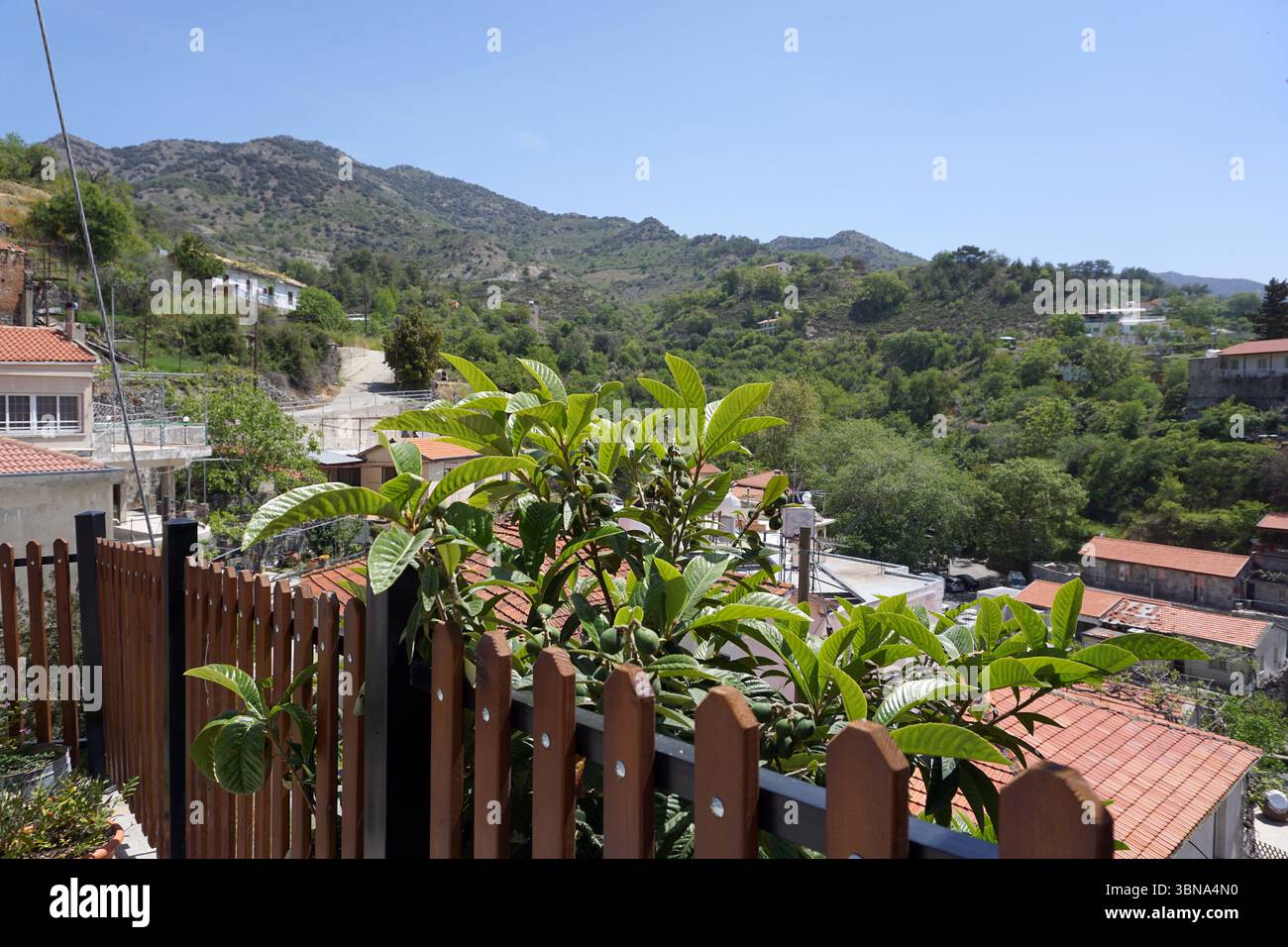 Eine ruhige Berglandschaft mit einem klaren blauen Himmel. Die Berge sind von üppiger grüner Vegetation bedeckt und das Gelände ist hauptsächlich felsig und hügelig. Im Vordergrund steht ein hölzerner Zaun mit einem schwarzen Metallgeländer, auf dem ein kleiner Baum oder Büsche wächst. Hinter dem Zaun befindet sich eine kleine Siedlung im Tal mit Gebäuden mit roten Ziegeldächern. Die Siedlung ist von einer Mischung aus Bäumen und Vegetation umgeben, und in der Mitte ist eine asphaltierte Straße oder ein gepflasterter Pfad zu sehen. Das Bild wird von einem hohen Aussichtspunkt aufgenommen und bietet einen Panoramablick auf die Siedlung und den umliegenden Moun Stockfoto