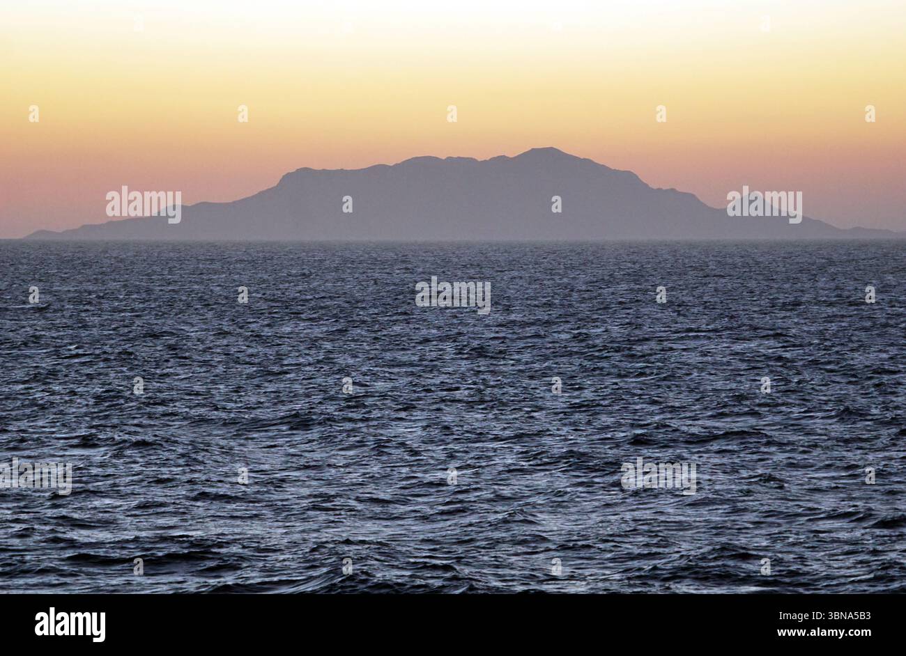 Griechische Inseln zwischen Zypern und Athen. Ein ruhiger Sonnenuntergang über einer riesigen Weite des dunkelblauen Ozeans. Der Himmel ist in Orange- und Rosa-Tönen gemalt, während die Sonne hinter einer Bergkette in der Ferne untergeht. Die Gebirgslandschaft, die sich gegen den Himmel schmiegt, scheint eine große, möglicherweise vulkanische Formation zu sein. Die Oberfläche des Ozeans wird von kleinen Wellen gestört, was der ansonsten ruhigen Szene ein Gefühl von Bewegung verleiht. Die Horizontlinie ist deutlich sichtbar und trennt den Ozean vom Himmel. Das Auge und die Phantasie eines Künstlers Stockfoto