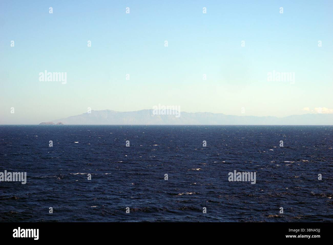 Griechische Inseln zwischen Zypern und Athen. Ein riesiger, tiefblauer Ozean, der sich bis zum Horizont erstreckt. Der Himmel ist klar, hellblau, mit ein paar Wolken am Horizont. In der Ferne erhebt sich eine Reihe von Bergen, vor allem braun und braun, am Horizont. Die Berge scheinen eine Mischung aus Felsen und möglicherweise etwas Vegetation zu sein. Das Bild wird von einem hohen Aussichtspunkt aufgenommen und bietet einen Panoramablick auf das Meer und die fernen Berge. Das Auge und die Phantasie eines Künstlers Stockfoto