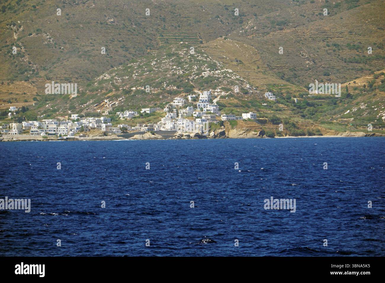 Griechische Inseln zwischen Zypern und Athen., Eine malerische Stadt auf einem Hügel mit weißen Gebäuden, die über die Landschaft verstreut sind. Die Stadt liegt auf einem Hügel, der zum tiefblauen Meer hin abfällt und einen markanten Kontrast zwischen den künstlichen Strukturen und der natürlichen Umgebung schafft. Der Hügel ist mit Vegetation bedeckt, einschließlich Bäumen und Sträuchern, und scheint eine Mischung aus Vegetation und möglicherweise einigen Kulturflächen zu sein. Das Meer hat eine tiefblaue Farbe, mit kleinen Wellen, die sanft gegen das Ufer plätschern. Die Perspektive des Bildes ist vom Meer aus, in Richtung der Stadt, Stockfoto