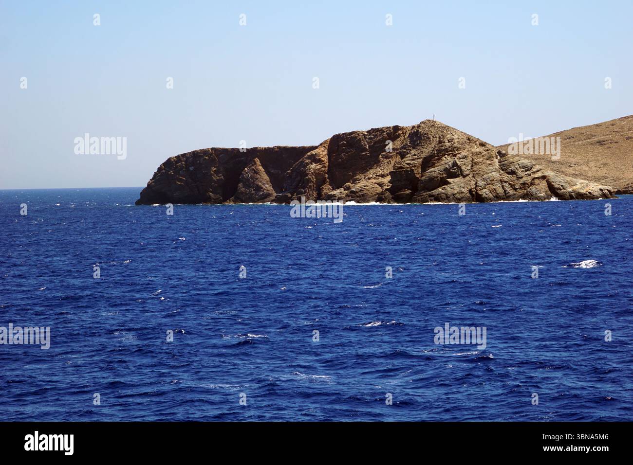 Griechische Inseln zwischen Zypern und Athen. Eine felsige Insel inmitten eines riesigen, tiefblauen Ozeans. Die Insel besteht hauptsächlich aus dunkelbraunen und grauen Felsen, wobei einige hellere Felsen über ihre Oberfläche verstreut sind. Das Meer, das die Insel umgibt, ist relativ ruhig, mit kleinen Wellen, die sanft gegen das felsige Ufer plätschern. Der Himmel darüber ist klar, hellblau. Ein kleines, dunkles Objekt, möglicherweise ein Leuchtturm, ist auf der Spitze der Insel zu sehen. Die Horizontlinie ist in der Entfernung deutlich sichtbar und trennt den Ozean vom Himmel. Das Auge und die Phantasie eines Künstlers Stockfoto