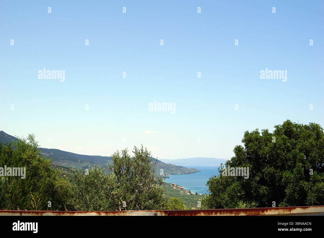 Eine ruhige Küstenlandschaft mit einem klaren blauen Himmel und einem ruhigen, hellblauen Meer. Das Meer erstreckt sich zum Horizont hin und trifft auf eine Reihe grüner Berge in der Ferne. Im Vordergrund befinden sich ein rustikaler Holzzaun und üppig grüne Bäume, die eine natürliche Grenze zur Szene bilden. Die Berge in der Ferne scheinen von Vegetation bedeckt zu sein, möglicherweise von Feldern oder Weinbergen. Das Bild wird von einem hohen Aussichtspunkt aufgenommen und bietet einen Panoramablick auf die Landschaft. Bildunterschrift in Form eines Künstlerauges und einer Fantasie., Nies, Griechenland Stockfoto