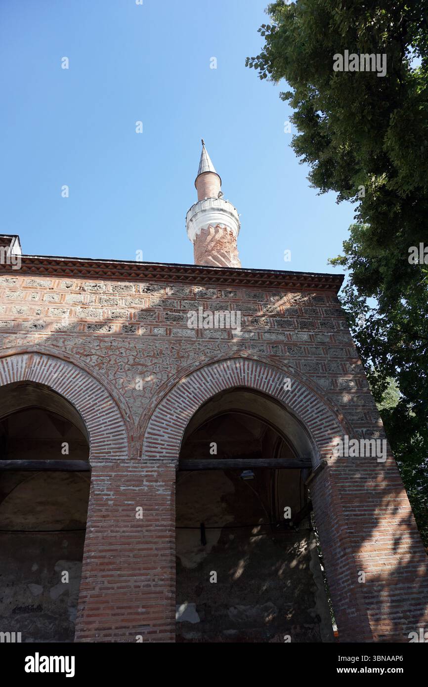 Plovdiw, Bulgariens zweitgrößte Stadt, ist eine der ältesten, durchgängig bewohnten Städte Europas mit einer über 8.000-jährigen Geschichte. Eingebettet am Maritsa River. Ein Teil eines roten Backsteingebäudes mit einem weißen Minarett und einem spitzen Dach vor einem klaren blauen Himmel. Das Gebäude verfügt über zwei bogenförmige Öffnungen an der Vorderseite, die als Teil eines überdachten Gehwegs oder Eingangs erscheinen. Das Mauerwerk hat ein strukturiertes Aussehen und das Minarett ist mit einer weißen Kuppel gekrönt. Die Perspektive des Bildes ist aus einem niedrigen Winkel, der auf das Minarett blickt und dessen Höhe und Pracht betont. Die Stockfoto