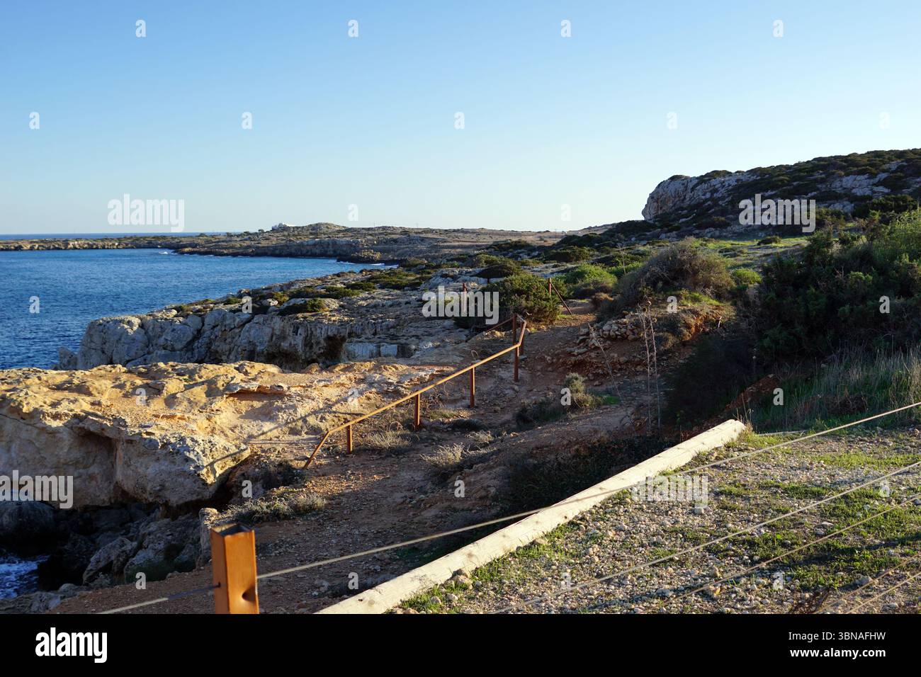 Eine felsige Küste mit einem klaren blauen Himmel. Ein hölzernes Geländer mit Metallgeländer verläuft am Rand der felsigen Klippe entlang und bietet Besuchern einen Weg, um die Gegend zu erkunden. Die Klippe ist mit einer Mischung aus grüner und brauner Vegetation bedeckt, und die felsige Oberfläche hat eine hellbraune oder beige Farbe. Der tiefblaue Ozean erstreckt sich bis zum Horizont und trifft auf den Himmel am Horizont. Das Bild wurde von einem hohen Aussichtspunkt aufgenommen und bietet einen Panoramablick auf die Küste und das Meer. Bildunterschrift in Form eines Künstlers mit Blick und Fantasie, Capo Greco, Zypern Stockfoto
