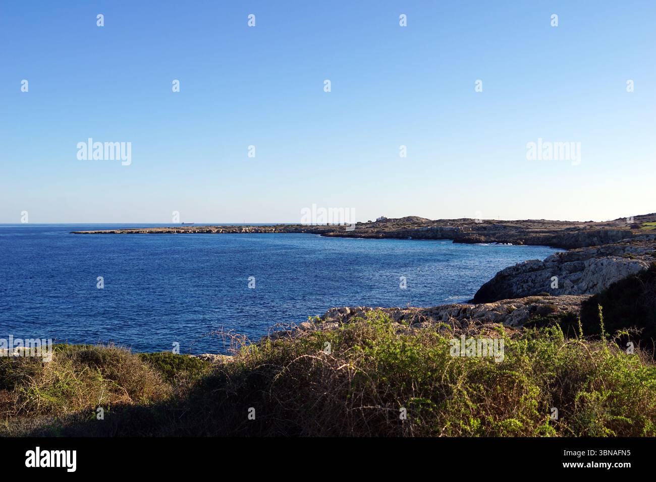 Eine ruhige Küstenlandschaft. Das tiefblaue Meer erstreckt sich bis zum Horizont und trifft auf den klaren, leuchtend blauen Himmel am oberen Rand des Rahmens. Der Vordergrund wird von einer felsigen Küste dominiert, mit einer Mischung aus hellen und dunkelgrauen/braunen Felsen und einigen Stellen grüner Vegetation. Die felsige Küste wird von einer kleinen, felsigen Insel in der Mittelweite begrenzt. Die Perspektive des Bildes ist von einem hohen Aussichtspunkt aus und bietet einen Panoramablick auf die Küste und die weite Weite des Meeres. Bildunterschrift in Form eines Künstlers mit Blick und Fantasie, Capo Greco, Zypern Stockfoto