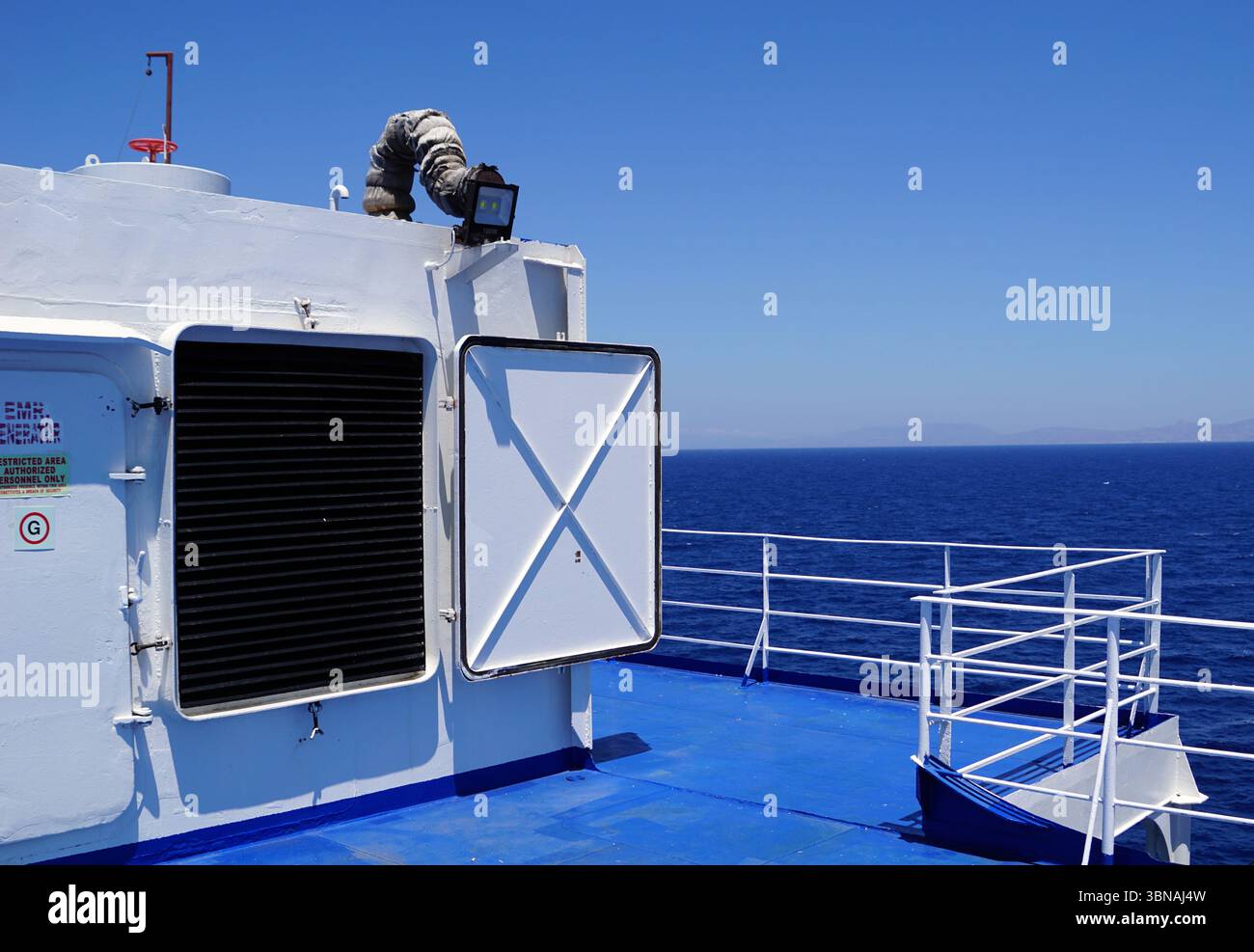 Griechische Inseln zwischen Zypern und Athen. Das Bild zeigt das Oberdeck einer Fähre, die in einem makellosen weißen Farbton gemalt ist. Das Deck verfügt über ein blaues Geländer und ein weißes Geländer, was einen Kontrast zur weißen Struktur bildet. Eine schwarze Lüftungsöffnung befindet sich auf der linken Seite des Decks und eine weiße Tür mit einem schwarzen Rolltor auf der rechten Seite. Ein rot-weiß gestreiftes Objekt, möglicherweise ein Rettungsschirm, ist auf dem Dach der Terrasse montiert. Auf dem Dach ist auch eine schwarze Entlüftung oder Klimaanlage zu sehen. Das Deck befindet sich auf einer blauen Oberfläche, wahrscheinlich auf einem Deck oder einer Plattform, und der riesigen Wasserfläche Stockfoto